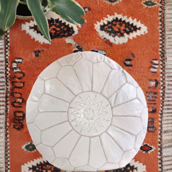 White Moroccan leather ottoman pouffe on patterned orange rug.